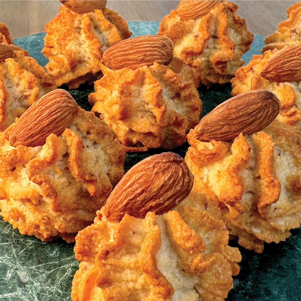 True Delicious Italian Almond Cookies feature a golden-brown, rough texture and are topped with whole almonds. Arranged on a dark green marbled surface, these classic Sicilian cookies are naturally gluten-free.