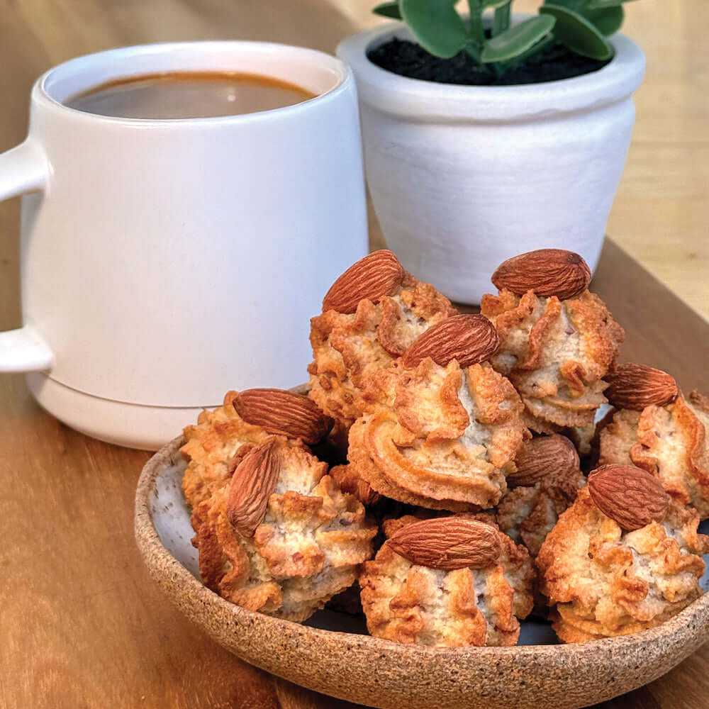 A ceramic mug of coffee sits beside a small potted plant and a bowl of True Delicious Italian Almond Cookies, each gluten-free and topped with a whole almond, on a wooden surface.