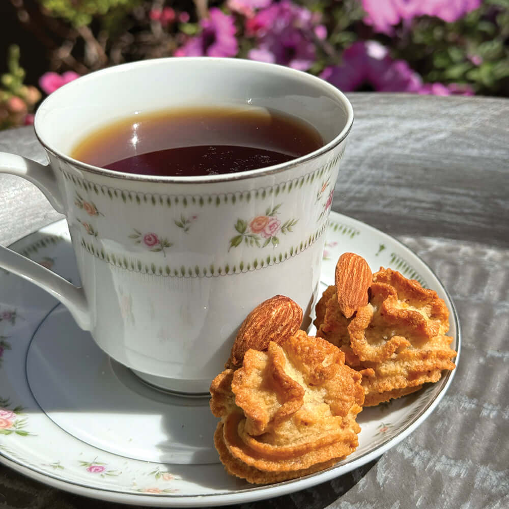 A floral teacup of black tea rests on a matching saucer beside two True Delicious Italian Almond Cookies, made with a traditional Sicilian recipe. Blurred pink flowers and greenery are visible in the background.