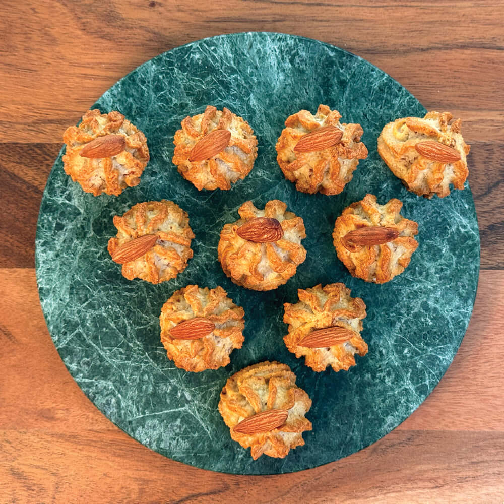 Eleven True Delicious Italian Almond Cookies—small, round treats with ridged edges and whole almonds on top—are arranged on a green marble plate atop wood, featuring a classic gluten-free Sicilian recipe.