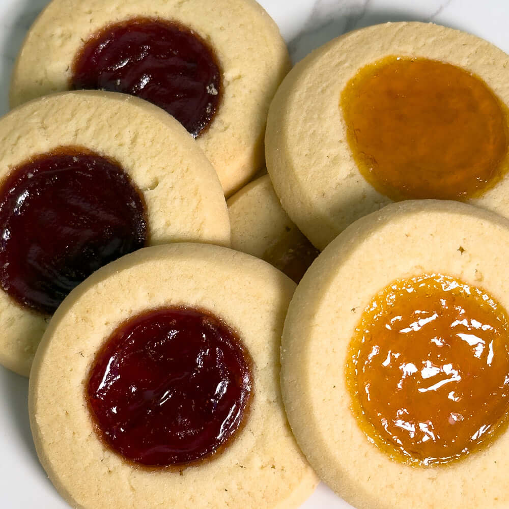 A close-up of True Delicious Jam Cookies Apricot Raspberry—six buttery shortbread cookies, half with raspberry and half with apricot jam—arranged on a white plate.