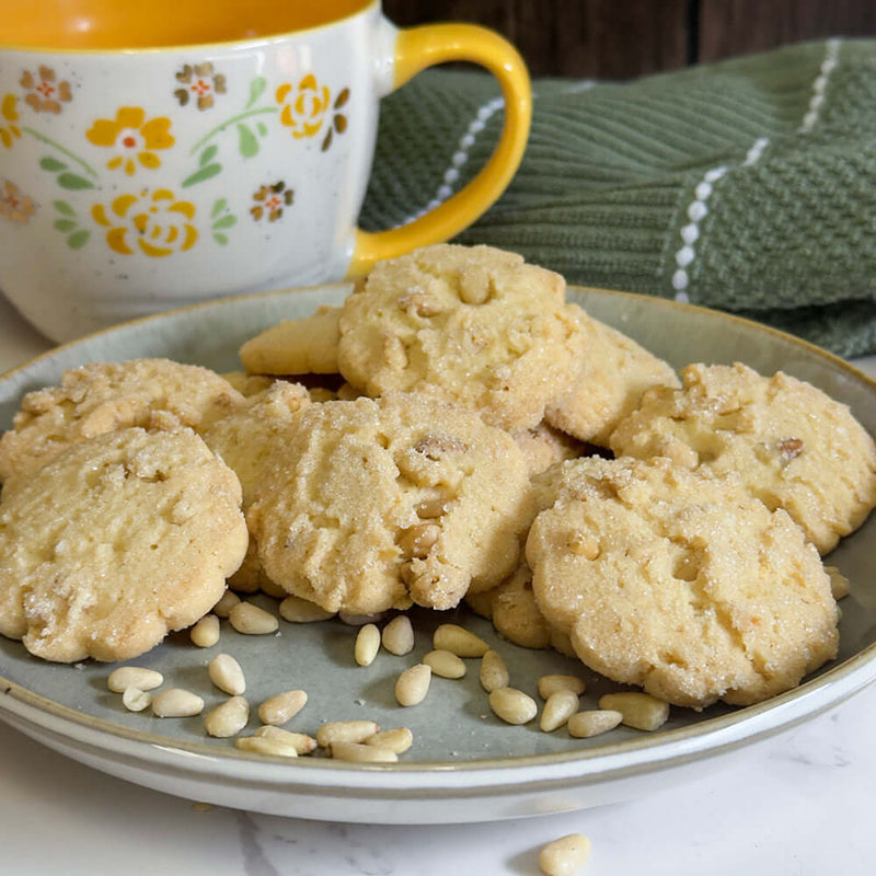 True Delicious Pine Nut Tea Cookies are arranged on a marble surface with toasted pine nuts nearby. A floral mug and a green striped cloth add charm in the background.