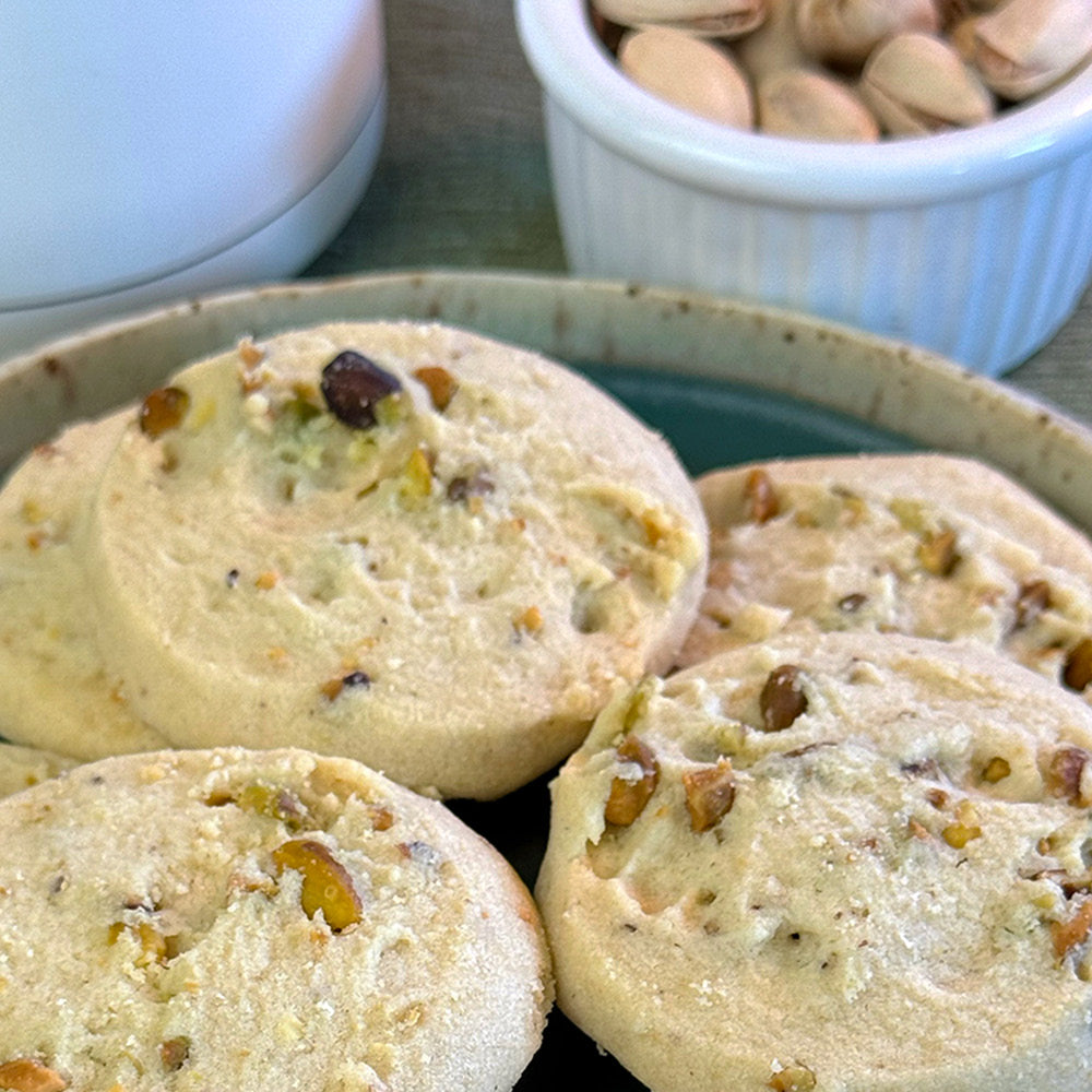 A close-up of True Delicious Biscotti Pistachio Shortbread Cookies on a plate, featuring crushed pistachios in the dough. In the background are a white bowl filled with unshelled pistachios and part of a white container.