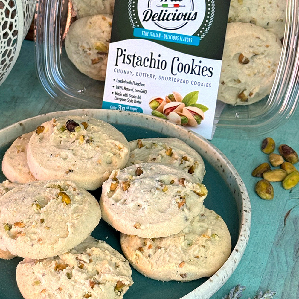 A plate of Pistachio Shortbread Cookies by True Delicious Biscotti is in front of an open container labeled Pistachio Cookies, with whole pistachios scattered on a light blue surface.