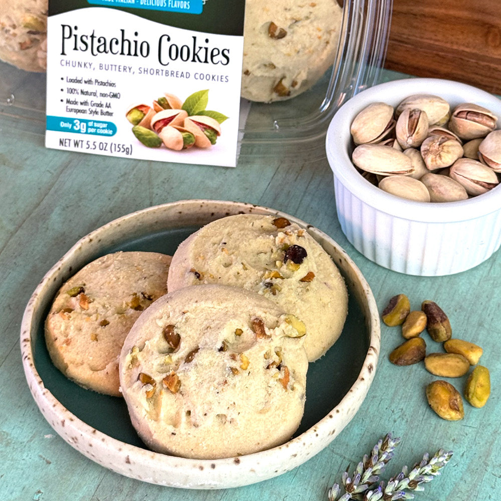 A ceramic plate with three Pistachio Shortbread Cookies from True Delicious Biscotti sits on a blue surface, accompanied by a small bowl of pistachios, loose pistachios, and lavender sprigs. A package labeled Pistachio Shortbread Cookies is in the background.
