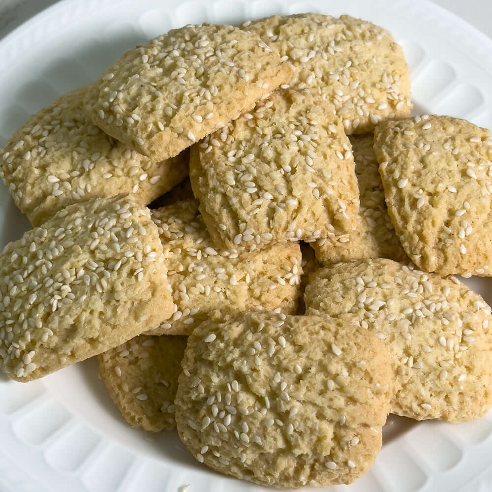 A white plate holds square-shaped, golden Reginelle Sesame Cookies by True Delicious—Sicilian shortbread topped with toasted white sesame seeds—arranged in a slightly overlapping pile.