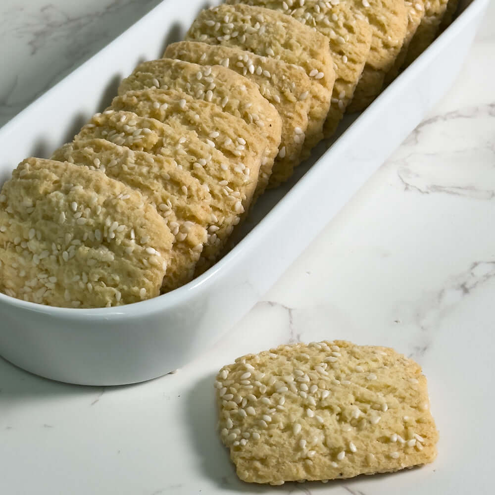 A row of True Delicious Reginelle Sesame Cookies are arranged in a white dish, each golden and topped with toasted sesame seeds. One cookie sits on the white marble surface beside the dish.