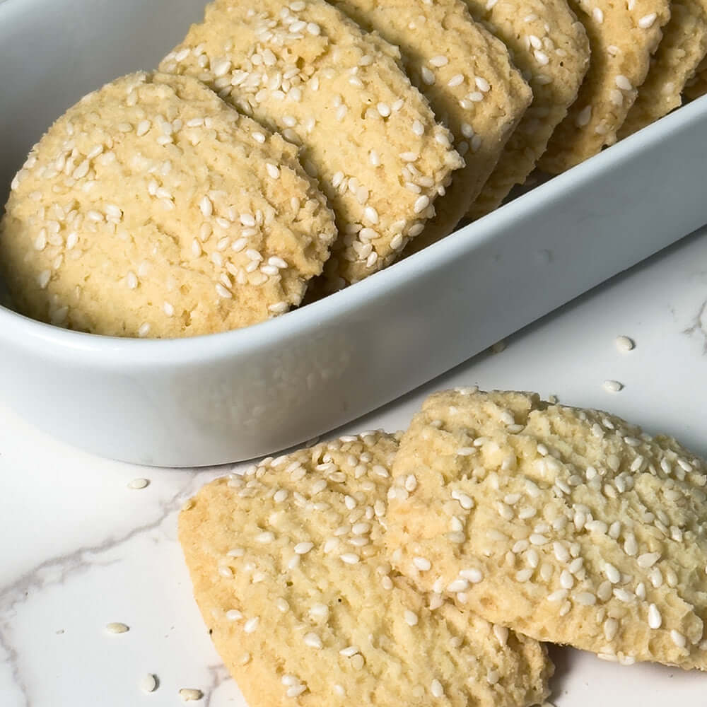 True Delicious Reginelle Sesame Cookies, a Sicilian shortbread topped with toasted sesame seeds, are displayed in a white rectangular dish with a few on the marble beside it.