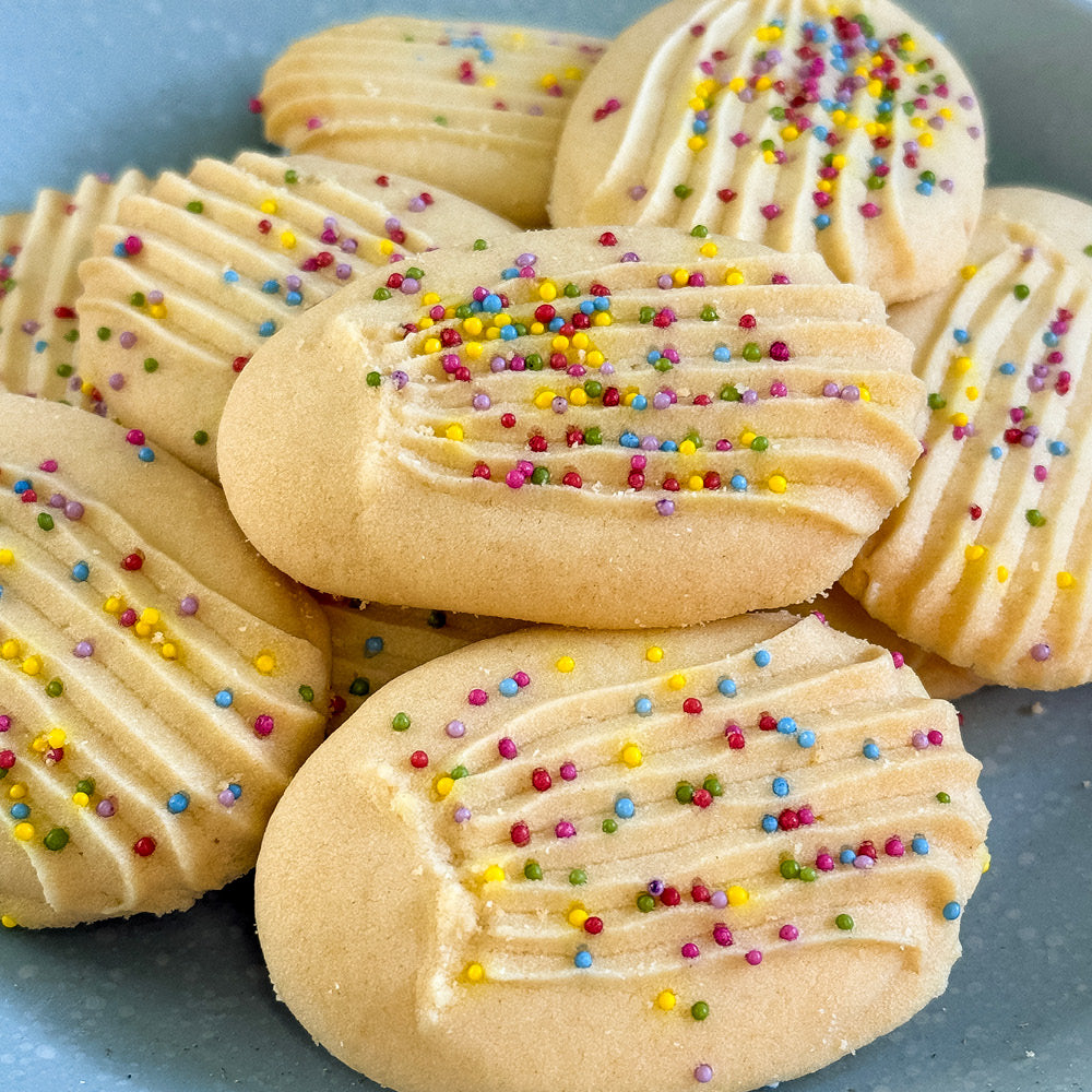 True Delicious Sprinkle Cookies are oval-shaped Italian-American treats with a ridged top, topped with naturally colored sprinkles and arranged in a slightly overlapping pile, making them a perfect choice for any occasion.