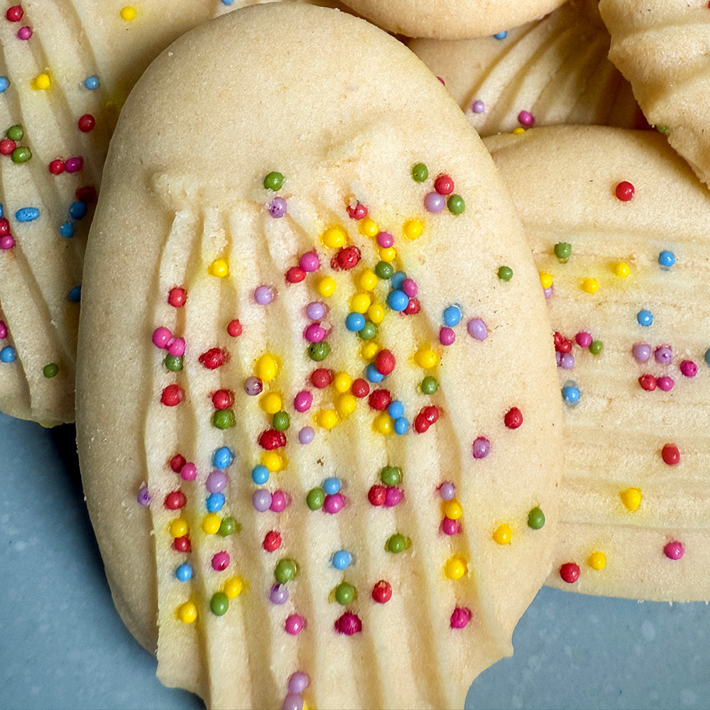 A close-up of True Delicious Sprinkle Cookies with a ridged pattern, topped with naturally colored pink, yellow, green, purple, and red sprinkles. The cookies are stacked on a light surface.