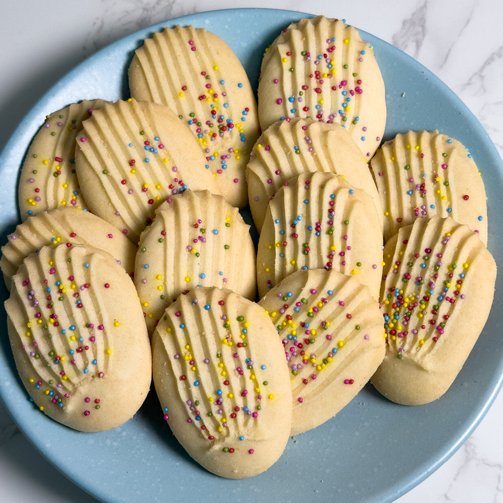 A blue plate displays True Delicious Sprinkle Cookies—oval-shaped Italian-American treats with naturally colored sprinkles and ridged lines—resting on a white marble surface.