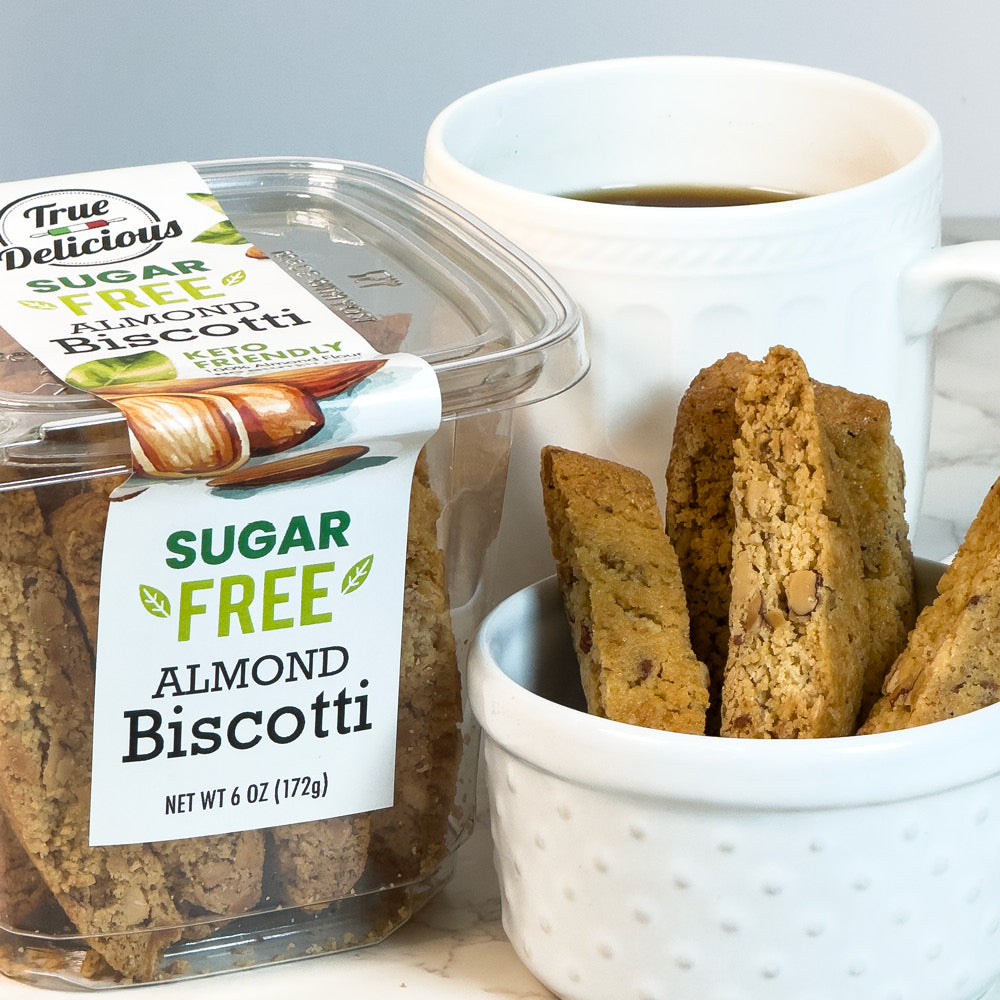 A container of True Delicious Sugar Free Almond Biscotti sits beside a white coffee mug and a bowl of biscotti pieces, with the packaging clearly showing the Sugar Free Almond Biscotti label.