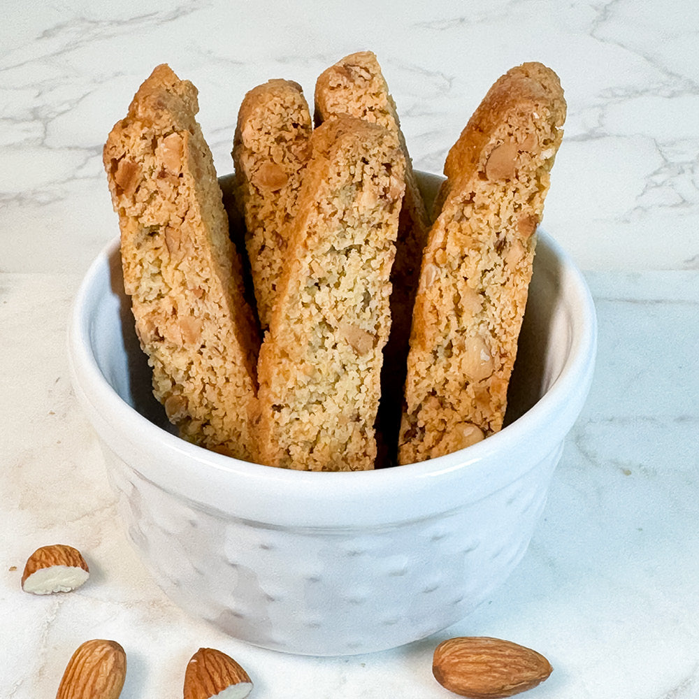 A white bowl holds several True Delicious Sugar Free Almond Biscotti, with whole and halved almonds scattered nearby on a white marble surface.