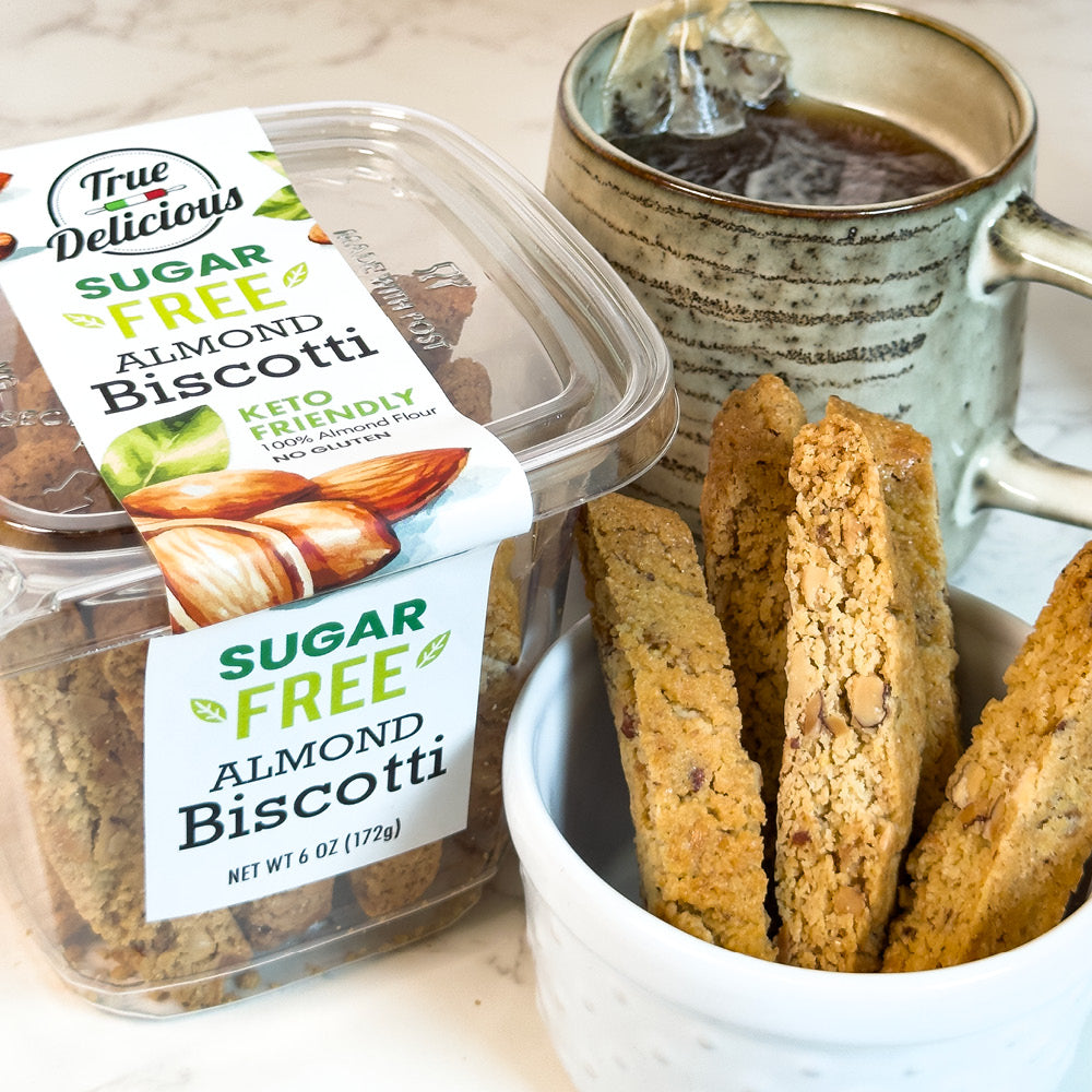 A container of True Delicious Sugar Free Almond Biscotti is beside a mug of steaming tea and a bowl with several keto biscotti pieces on a light surface.