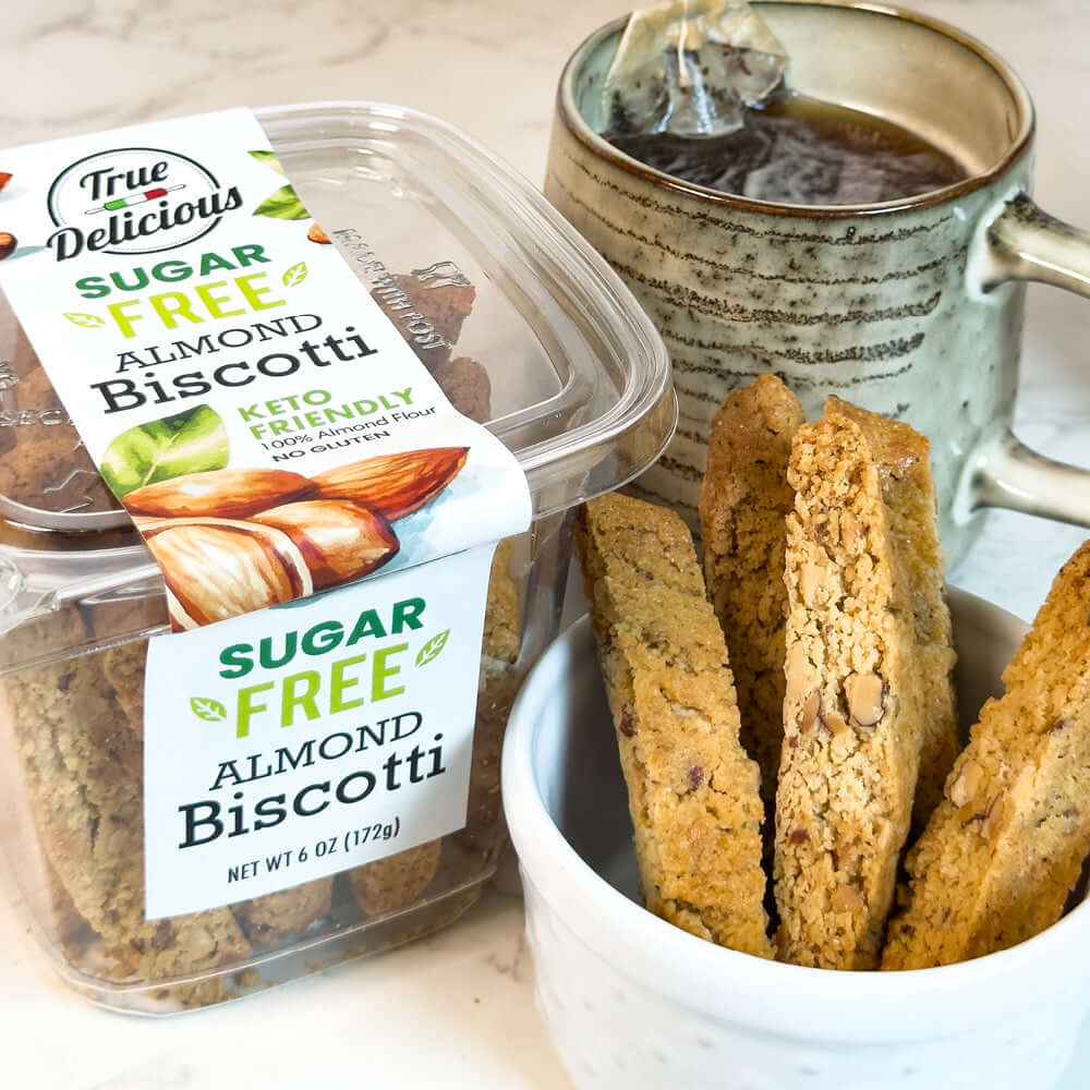 A container of True Delicious Sugar Free Almond Biscotti beside a mug of steaming tea and a bowl of biscotti pieces.