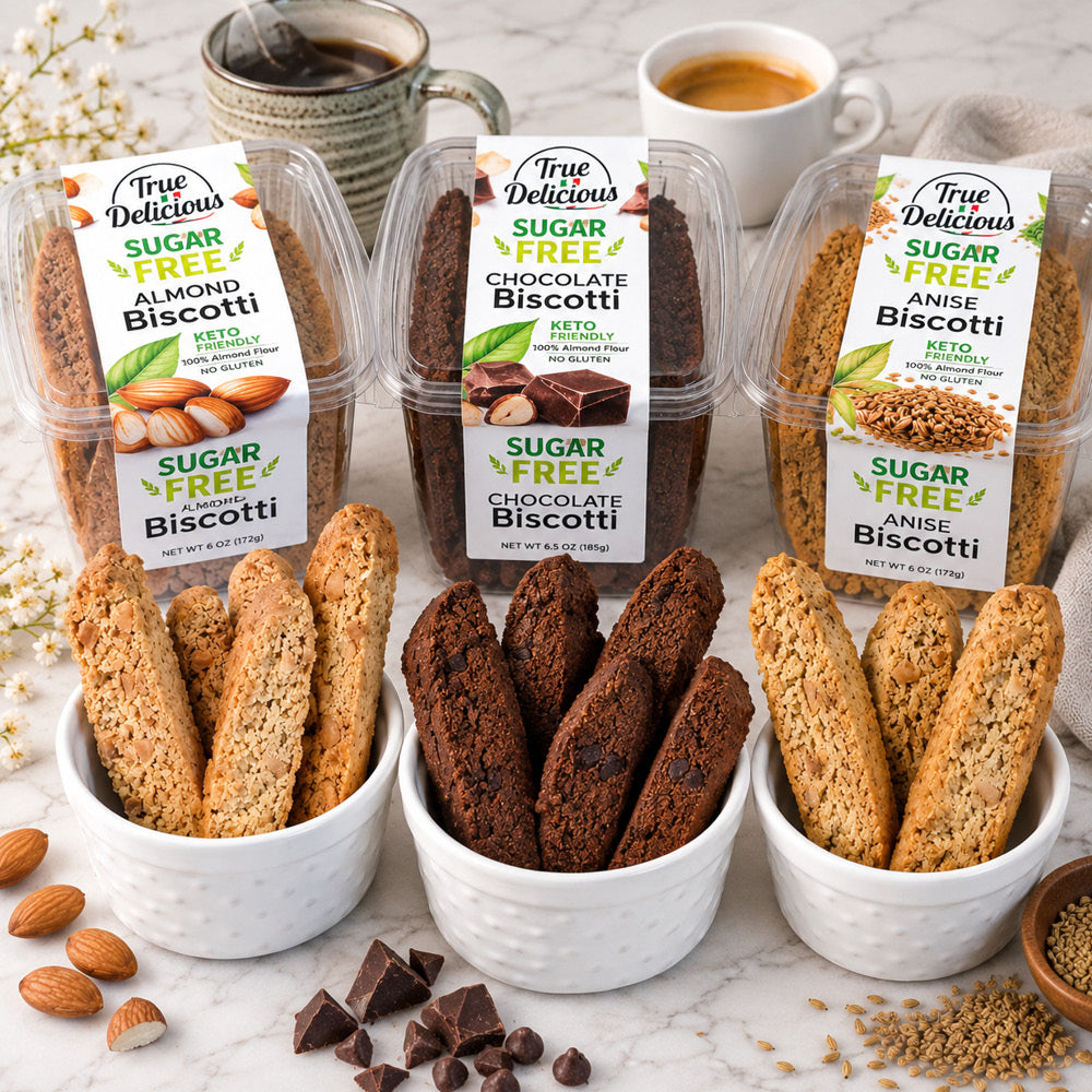 Six containers of True Delicious Sugar-Free Biscotti Variety 6-Pack are displayed along with matching almond flour biscotti in bowls, accompanied by coffee cups, almonds, chocolate chips, and white flowers on a table for a delectable keto-friendly treat.