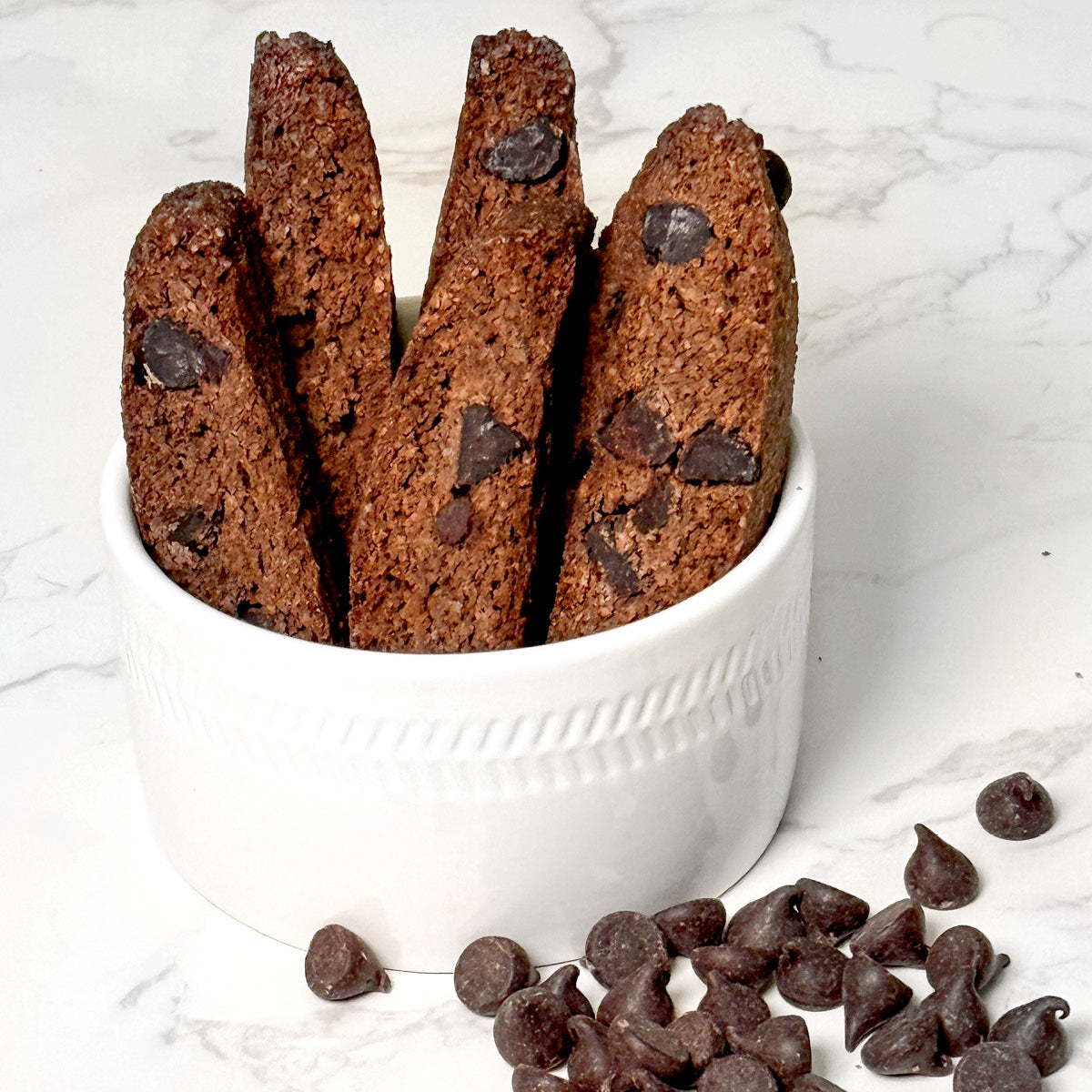 Four True Delicious Sugar Free Chocolate Biscotti, studded with chocolate chips, are arranged upright in a white bowl, with extra chocolate chips scattered on the white marble surface beside the bowl.