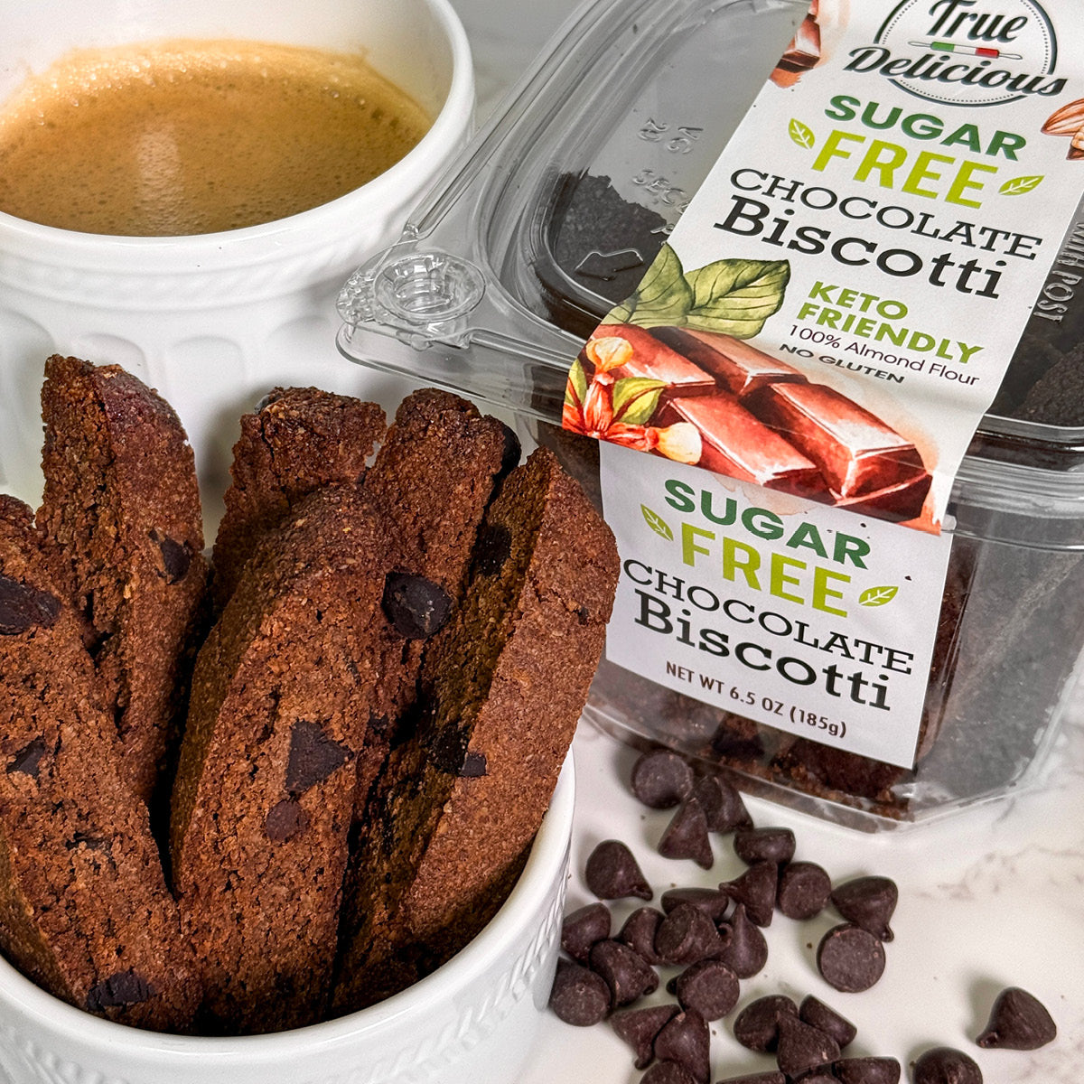 A cup of coffee sits next to a bowl with True Delicious Sugar Free Chocolate Biscotti, while chocolate chips are scattered on the white surface.